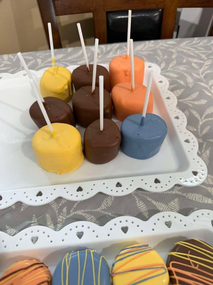 Cake pops in mustard, chocolate, orange and slate blue on a white tray, with drizzled dipped Oreos alongside.
