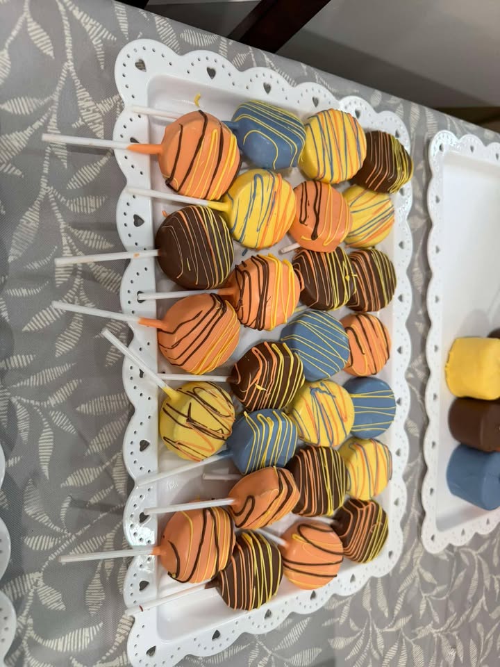 Dipped Oreos and cake pops drizzled in orange, blue and yellow on a scalloped white tray.