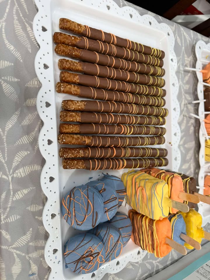 Chocolate-dipped pretzel rods beside blue and orange drizzled Oreos and cake pops.