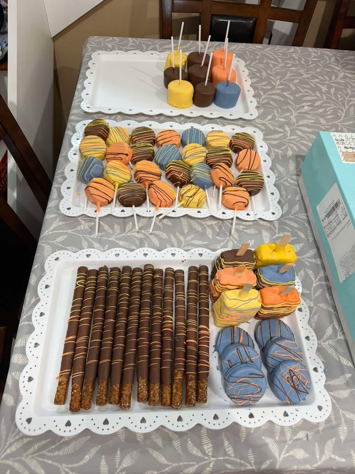 A tiered spread of dipped Oreos, chocolate pretzel rods and cake pops in blue, orange and yellow.
