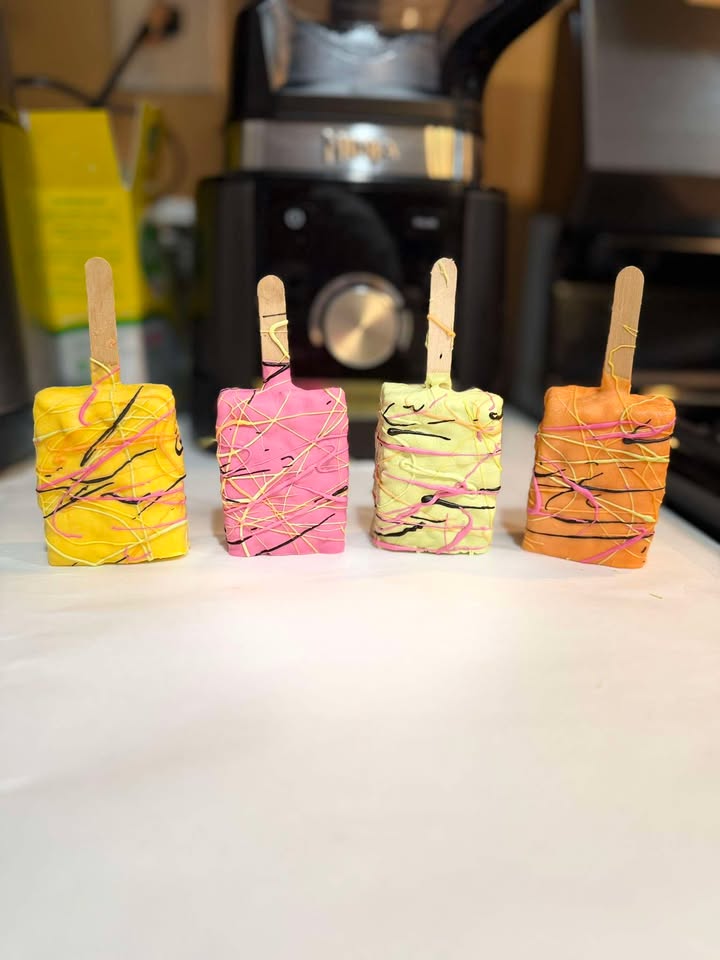 Yellow and pink drizzled cakesicles standing upright on a white counter.